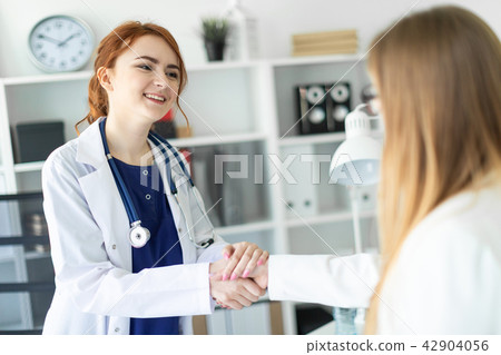 A beautiful young girl in a white coat is standing near the desk in the office and shaking hands 42904056