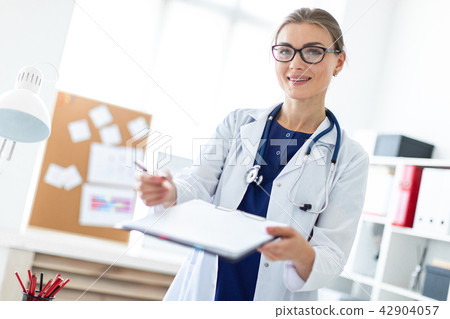 A young girl in a white coat is standing near a table in her office and holding a tablet and a pen A young girl in a white coat is standing near a table in her office and holding a tablet and a pen 42904057