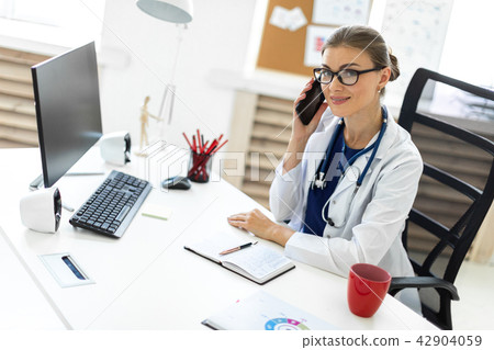 A young girl in a white robe is sitting at a table in the office and is talking on the phone. A A young girl in a white robe is sitting at a table in the office and is talking on the phone. A 42904059