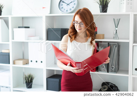 A beautiful young girl stands near a stack in the office and holds a folder with documents in her A beautiful young girl stands near a stack in the office and holds a folder with documents in her 42904062
