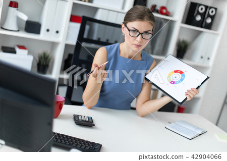 A young girl sits at a table in the office and holds a pen and a document with a diagram. A young girl sits at a table in the office and holds a pen and a document with a diagram. 42904066