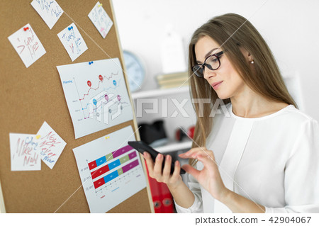 A young girl stands near a board with stickers and holds a phone in her hands. A young girl stands near a board with stickers and holds a phone in her hands. 42904067