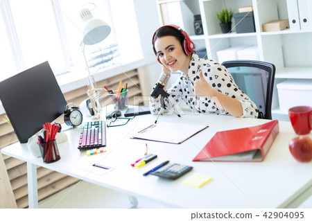 A beautiful young girl sits in headphones and with a microphone at the table in the office and shows 42904095