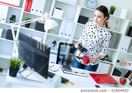 A young girl stands in the office near the table and pours coffee from the coffee pot into a red cup 42904097