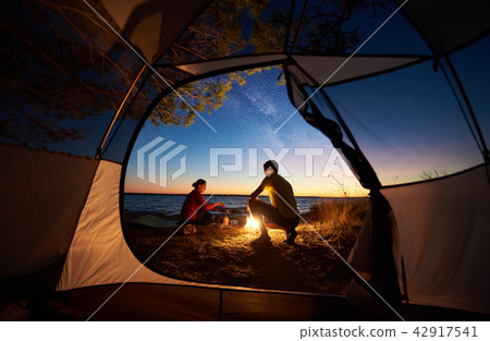 Young couple man and woman having rest at tourist tent and burning campfire on sea shore near forest Young couple man and woman having rest at tourist tent and burning campfire on sea shore near forest 42917541
