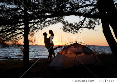Young couple man and woman having rest at tourist tent and burning campfire on sea shore near forest Young couple man and woman having rest at tourist tent and burning campfire on sea shore near forest 42917542
