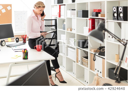 A young blond girl crouched on the desk in the office, talking on the phone and holding a notebook 42924648