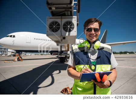 Airport worker looking at camera with smile while writing on clipboard Airport worker looking at camera with smile while writing on clipboard 42933616