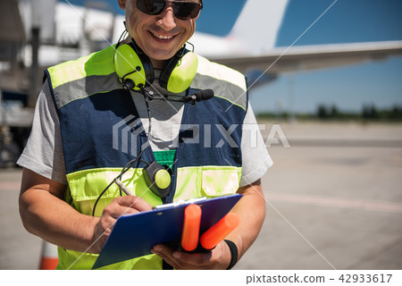 Cheerful airport worker looking at clipboard and writing information Cheerful airport worker looking at clipboard and writing information 42933617