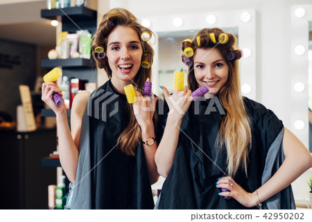 Two girls curling hair with curlers wearing cape having fun laughing putting rollers on fingers in Two girls curling hair with curlers wearing cape having fun laughing putting rollers on fingers in 42950202