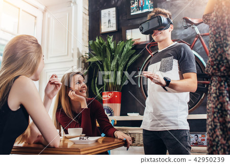 Group of young people having fun time together, cheerful guy wearing vr glasses in a cafe Group of young people having fun time together, cheerful guy wearing vr glasses in a cafe 42950239