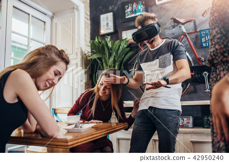 Young women laughing at their emotional male friend playing horror game in virtual reality goggles Young women laughing at their emotional male friend playing horror game in virtual reality goggles 42950240