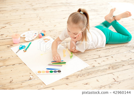 Top view of little blonde girl painting on big white paper while laying on the floor indoors Top view of little blonde girl painting on big white paper while laying on the floor indoors 42960144