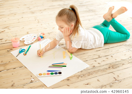 Top view of little blonde girl painting on big white paper while laying on the floor indoors Top view of little blonde girl painting on big white paper while laying on the floor indoors 42960145