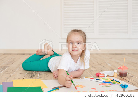 Little blonde girl painting on big white paper while laying on the floor indoors Little blonde girl painting on big white paper while laying on the floor indoors 42960195