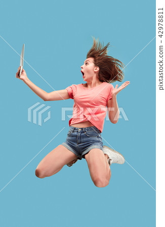 Image of young woman over blue background using laptop computer or tablet gadget while jumping. 42978811