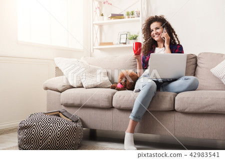 Smiling young woman with mobile, laptop and little dog at home 42983541