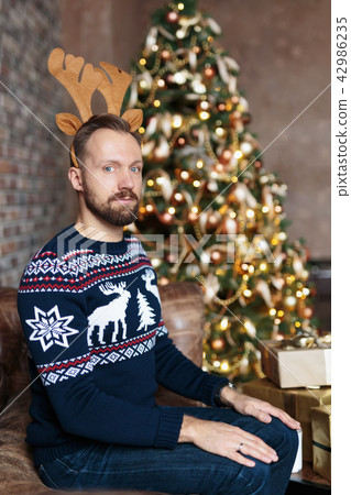 Young bearded man sitting on a sofa dressed Christmas jersey wearing antler deer headband 42986235