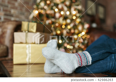 Adult person sitting on a sofa dressed Christmas socks. Golden lights on Christmas tree on the Adult person sitting on a sofa dressed Christmas socks. Golden lights on Christmas tree on the 42986263