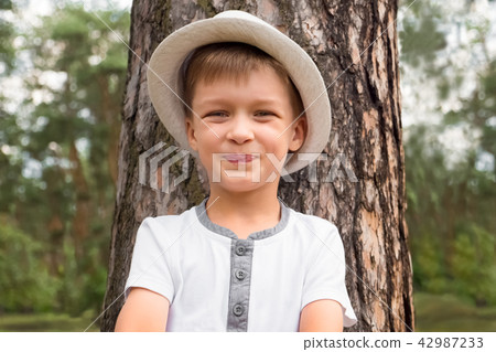 Happy boy in the hat is smiling near the tree trunk Happy boy in the hat is smiling near the tree trunk 42987233