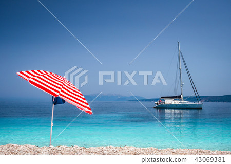 Bright red striped sun beach umbrella on pebble beach against turquoise blue shellow sea water and Bright red striped sun beach umbrella on pebble beach against turquoise blue shellow sea water and 43069381