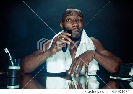 young man in bedroom sitting in front of the mirror scratching his beard 43083075