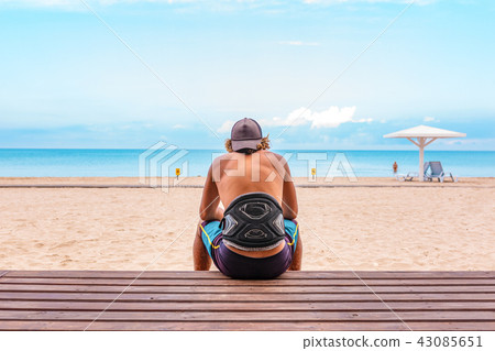 The surfer sits with his back to the camera on the terrace on the beach looking out into the The surfer sits with his back to the camera on the terrace on the beach looking out into the 43085651