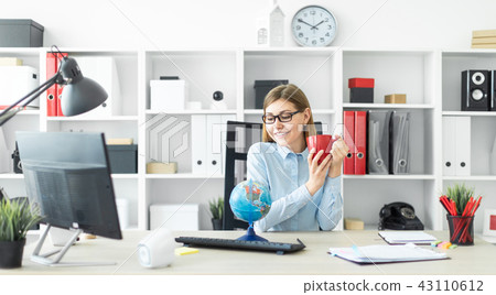 A young girl in glasses is sitting at a table, holding a red cup in her hands and looking at the 43110612