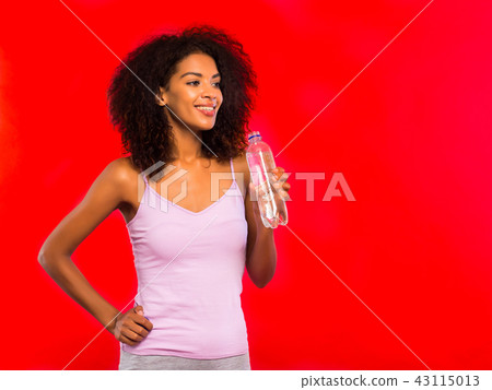 Attractive african-american sporty young woman drinking bottle of water on red wall background. Girl 43115013