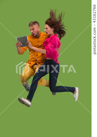 Jump of young couple over green studio background using laptop or tablet gadget while jumping. 43162766