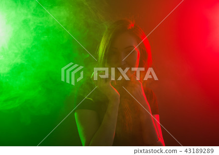 Portrait of young woman in neon red and green smoke with vape or e-cigarettes 43189289