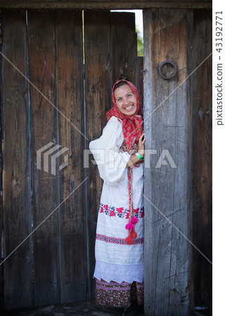 A woman in a Russian folk dress and a scarf standing at the wooden gate and smiling 43192771