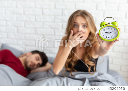 Late at work. Shocked girl holding alarm clock in bed 43204950