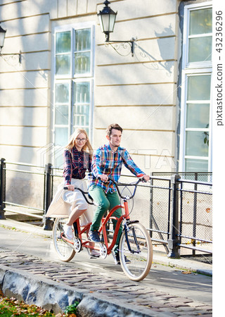 Young tourist couple, handsome man and pretty blond woman riding tandem bicycle along city street. 43236296