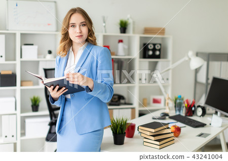 A beautiful young girl stands near a table in the office and holds an open book in her hands. 43241004