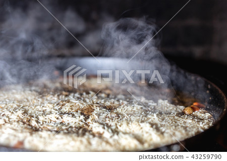 cooking Rice for pilaw or paella in iron cauldron over open fire. Smoke and steam. Tasty food cooking Rice for pilaw or paella in iron cauldron over open fire. Smoke and steam. Tasty food 43259790