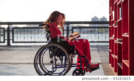 A young beautiful girl with red hair in a red dress is sitting in a wheelchair and reading a book 43261625