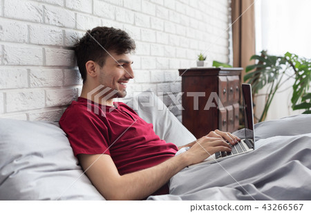 Happy handsome man using laptop in bed Happy handsome man using laptop in bed 43266567
