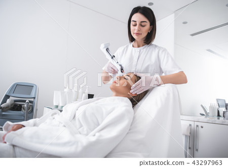 Young cosmetologist with automatic syringe gun making injection in girl forehead 43297963