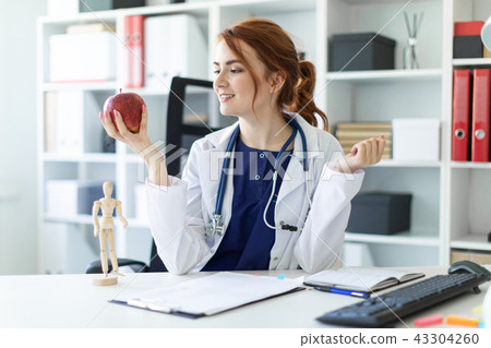 A beautiful young girl in a white robe is sitting at a table in the office and is holding an apple A beautiful young girl in a white robe is sitting at a table in the office and is holding an apple 43304260