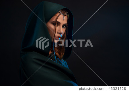 Beatiful elf princess wearing dark green cape looking at camera from darkness. 43321528