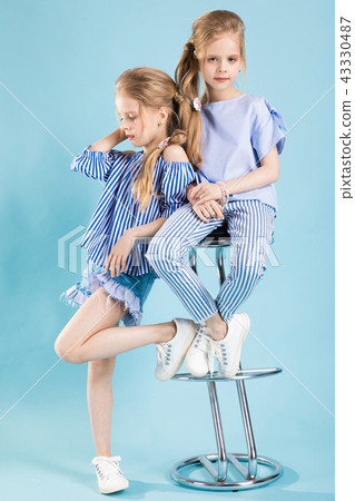 Girls twins in light blue clothes are posing near a bar stool on a blue background. Girls twins in light blue clothes are posing near a bar stool on a blue background. 43330487