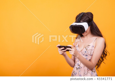 Woman wearing a futuristic looking virtual reality headset goggles in studio over yellow background Woman wearing a futuristic looking virtual reality headset goggles in studio over yellow background 43341771