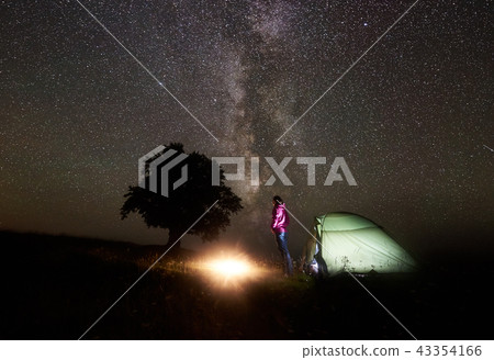 Young female tourist resting near illuminated tent, camping in mountains at night under starry sky 43354166
