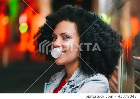 Young pretty woman with very curly afro hair inflates a bubble ball of white chewing gum at night Young pretty woman with very curly afro hair inflates a bubble ball of white chewing gum at night 43379048