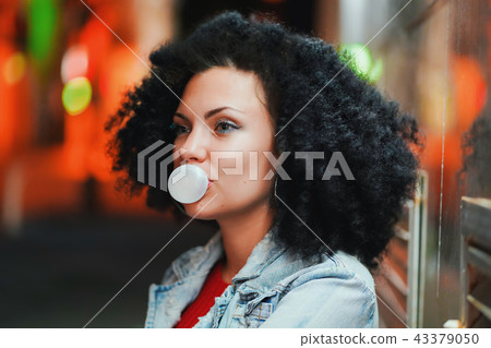 Young pretty woman with very curly afro hair inflates a bubble ball of white chewing gum at night Young pretty woman with very curly afro hair inflates a bubble ball of white chewing gum at night 43379050