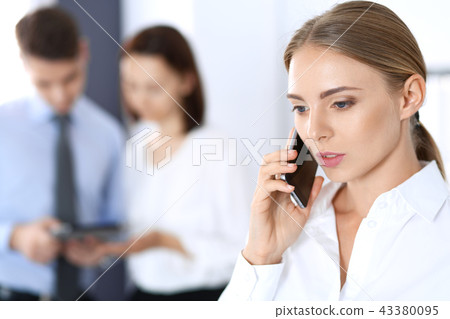 Serious blonde businesswoman talking by phone in the background of her colleagues in office Serious blonde businesswoman talking by phone in the background of her colleagues in office 43380095