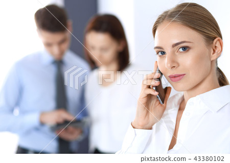 Serious blonde businesswoman talking by phone in the background of her colleagues in office Serious blonde businesswoman talking by phone in the background of her colleagues in office 43380102