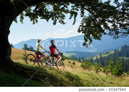 Cyclists, man and woman in helmets and full equipment, standing with bikes on grassy hill 43387944