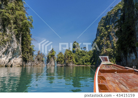 Wooden Thai traditional long-tail boat on a lake with mountains and rain forest in the background Wooden Thai traditional long-tail boat on a lake with mountains and rain forest in the background 43415159